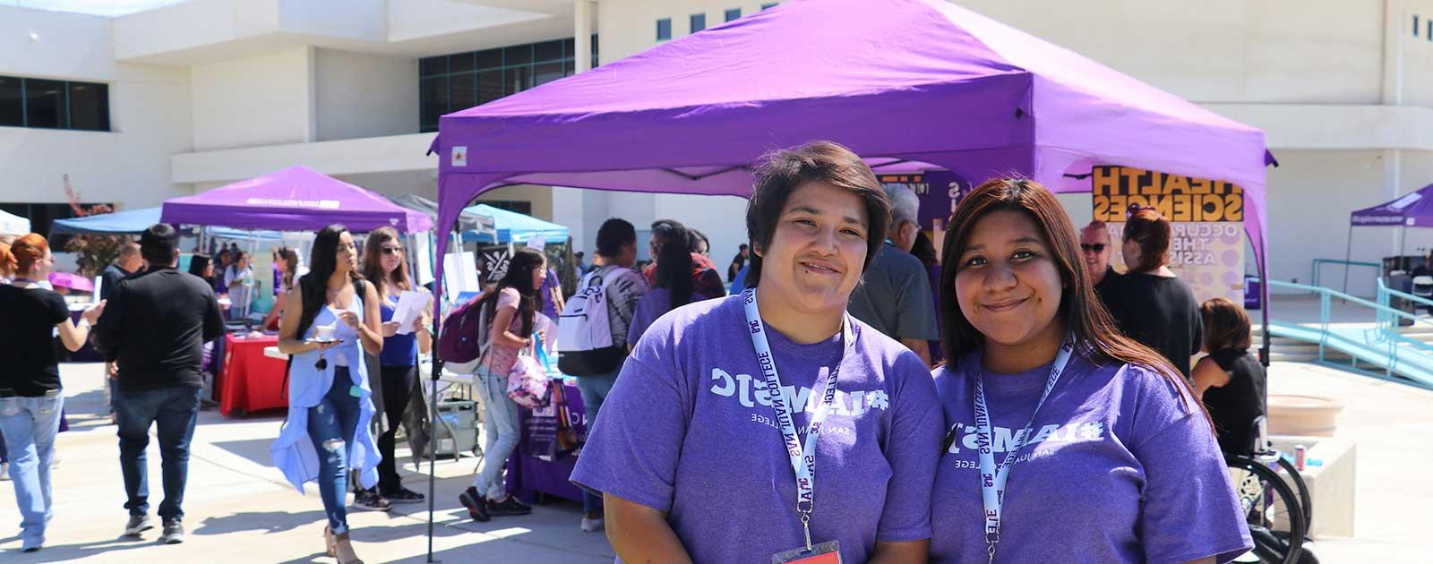 Two SJC students smiling at camera with SJC tents in background at outdoor event